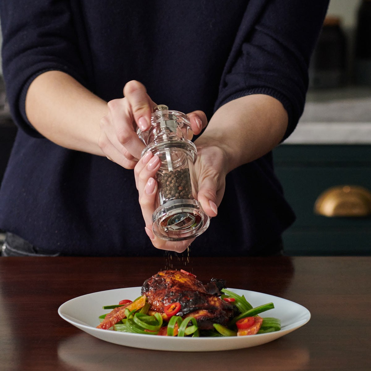 A person grinds black pepper onto a cooked piece of meat served with green beans and sliced tomatoes on a white plate, set on a dark wooden table.