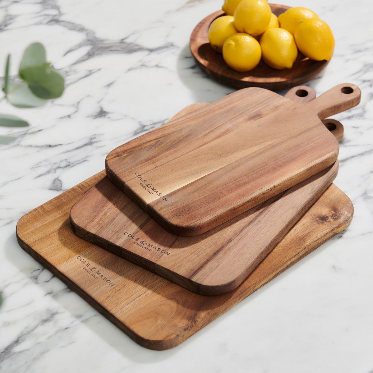 Three wooden cutting boards of varying sizes stacked on a marble countertop, with a wooden bowl of lemons and a sprig of greenery in the background.