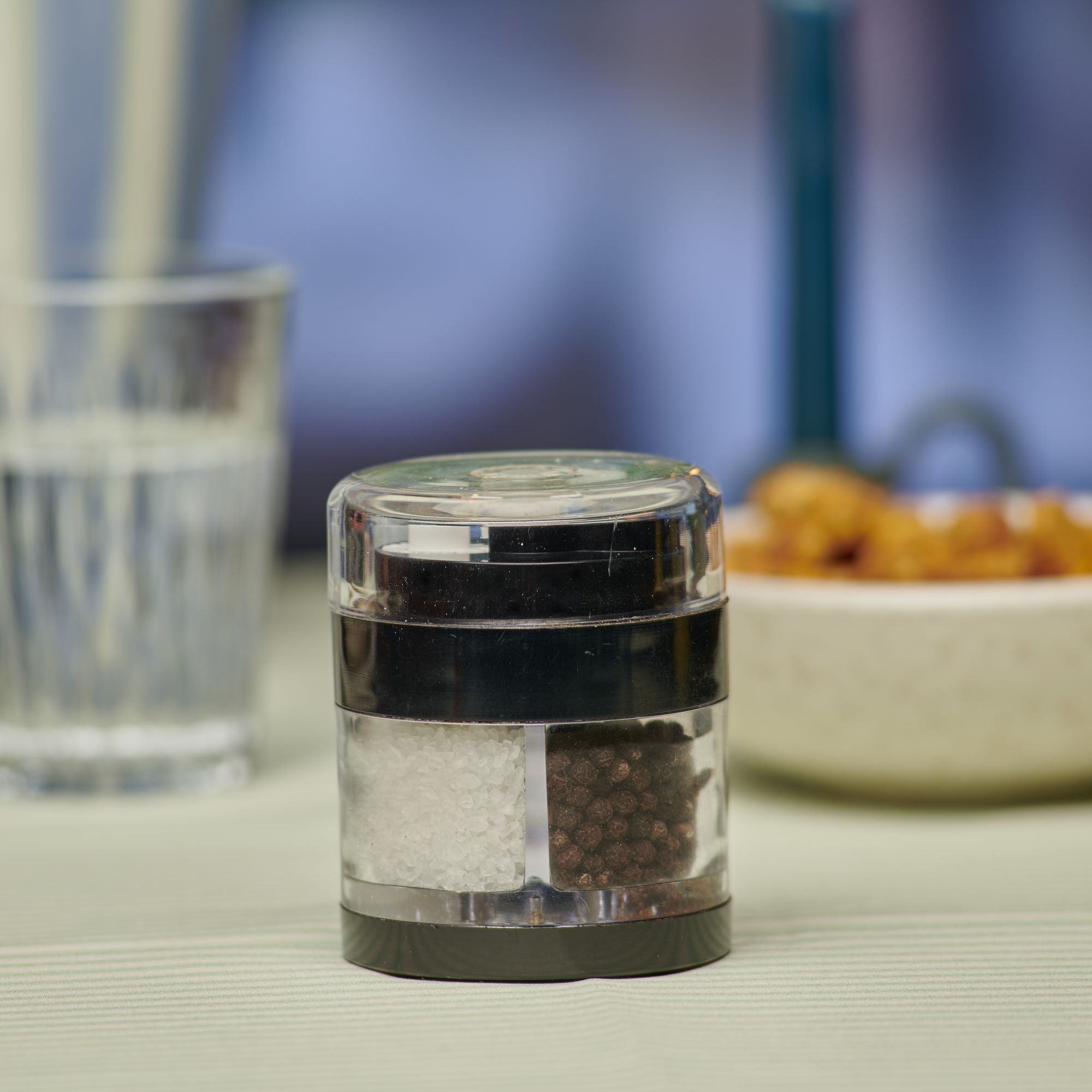 A dual-chamber grinder containing salt and black pepper sits on a light-colored table, with a glass of water and a bowl of food blurred in the background.