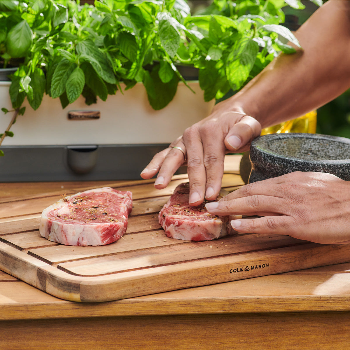A person seasons two raw steaks on a wooden cutting board outdoors, with fresh herbs and a mortar and pestle nearby.