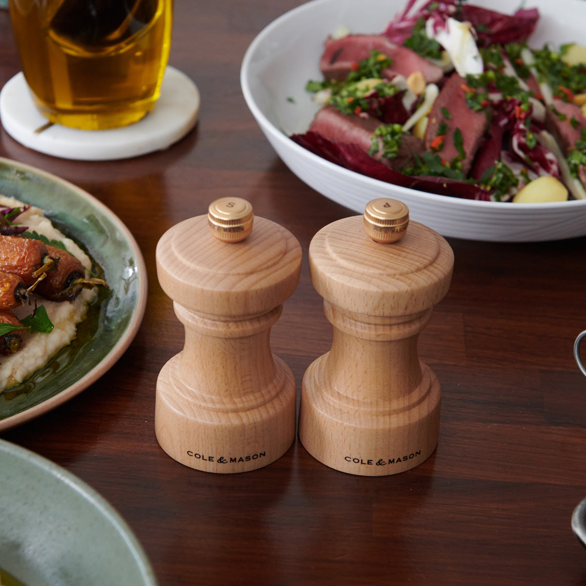 Two wooden Cole & Mason salt and pepper grinders sit on a wooden table, surrounded by plates of salad and various dishes.