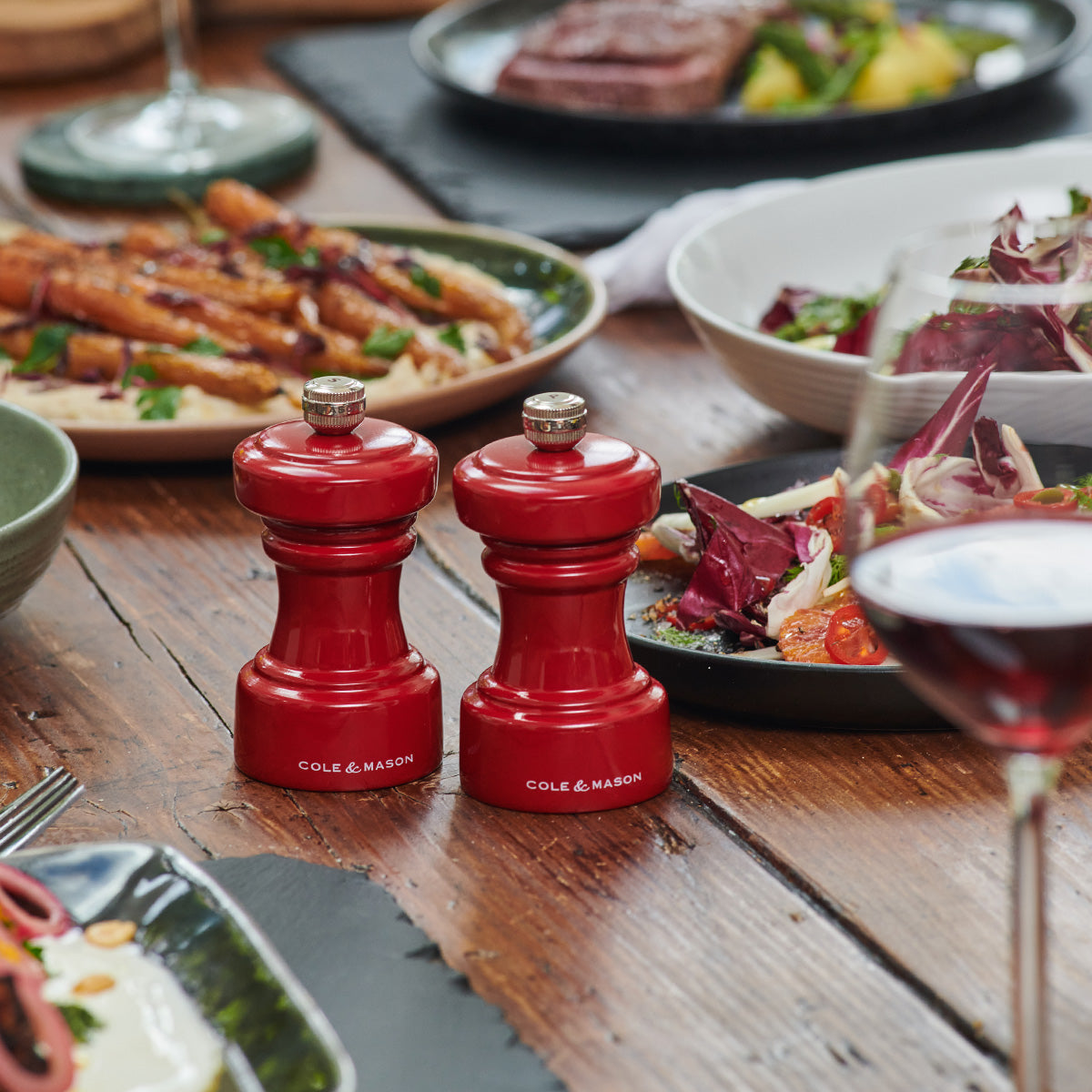 Two red Cole & Mason salt and pepper grinders sit on a wooden dining table surrounded by plates of food, salad, wine glasses, and cutlery.