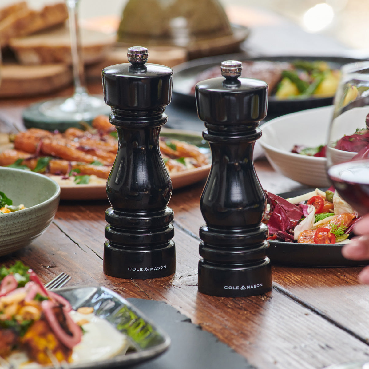Two black Cole & Mason salt and pepper grinders are placed on a wooden table, surrounded by plates of colorful salads, roasted dishes, and a hand holding a glass of red wine.