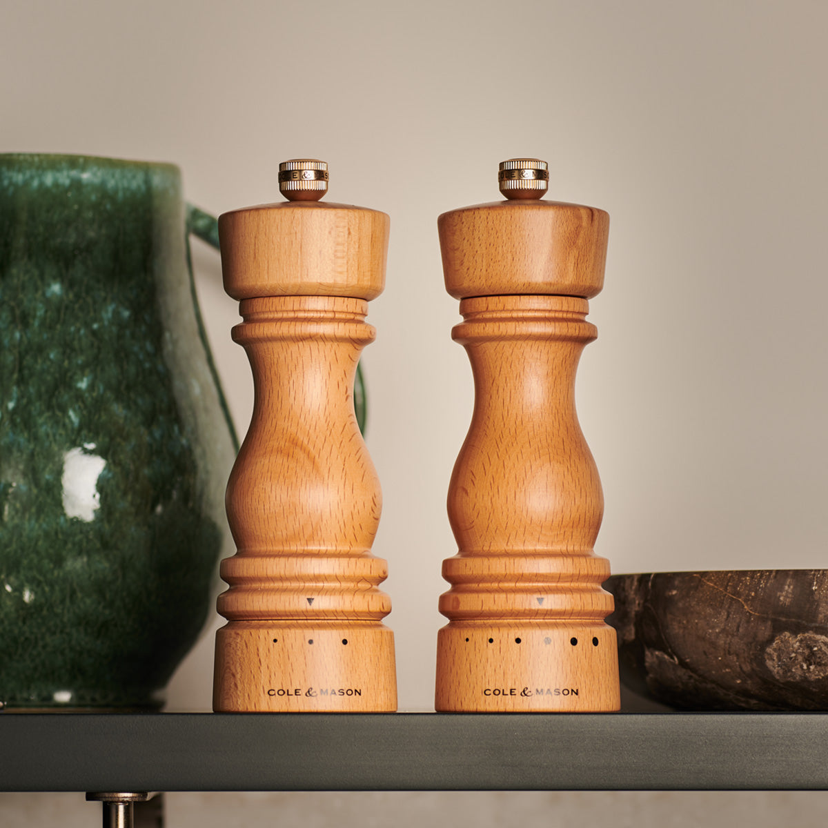 Two wooden Cole & Mason salt and pepper mills stand side by side on a shelf, next to a green ceramic pitcher and a wooden bowl, against a neutral background.