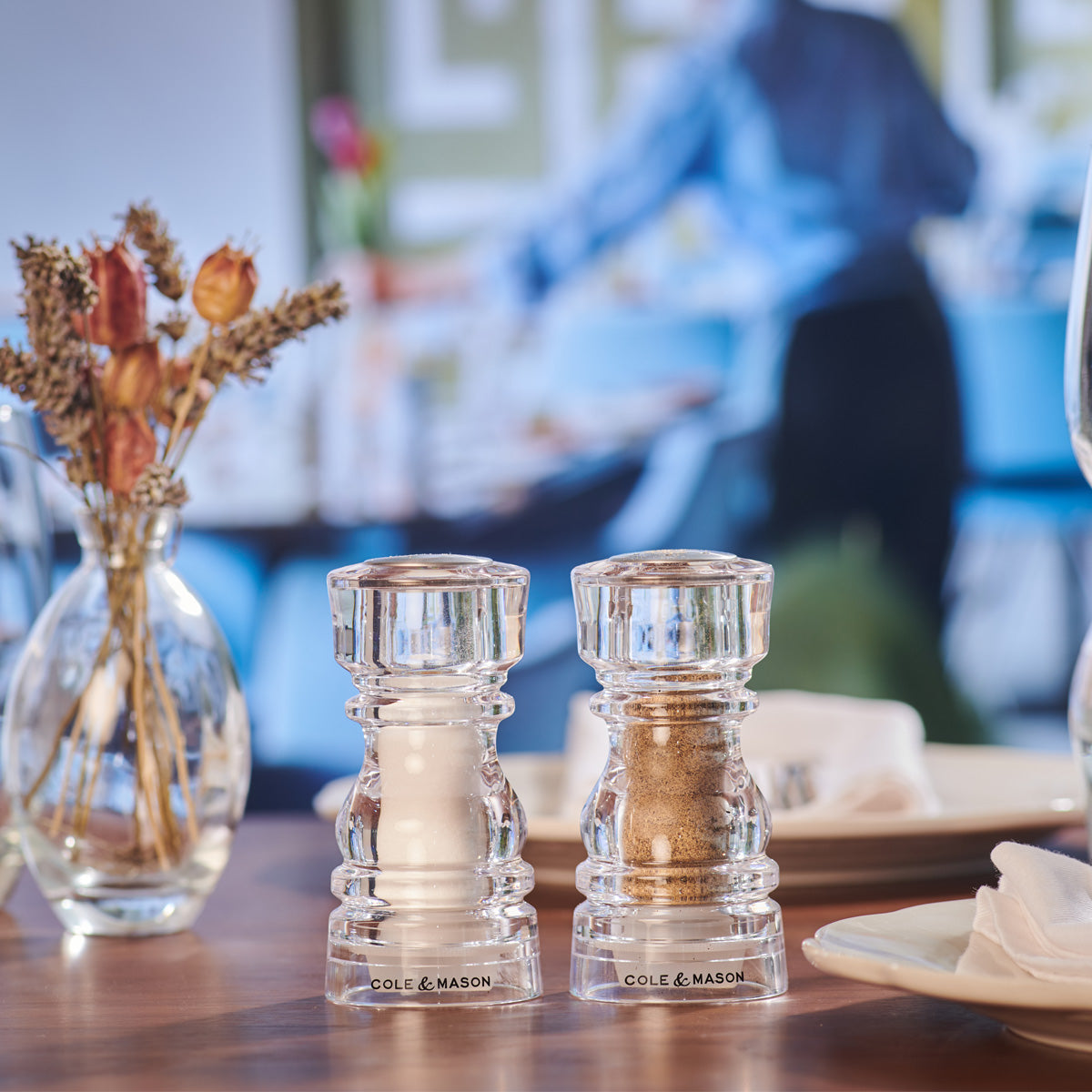 Two clear glass salt and pepper grinders sit on a set dining table. In the background, a blurry figure is seen standing, and a vase with dried flowers is also visible. The scene appears to be in a restaurant or café.