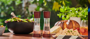 Two wooden salt and pepper mills sit on a table set with salad, bread, cheese, sandwiches, and a drink, with fresh greenery in the background and sunlight filtering through.