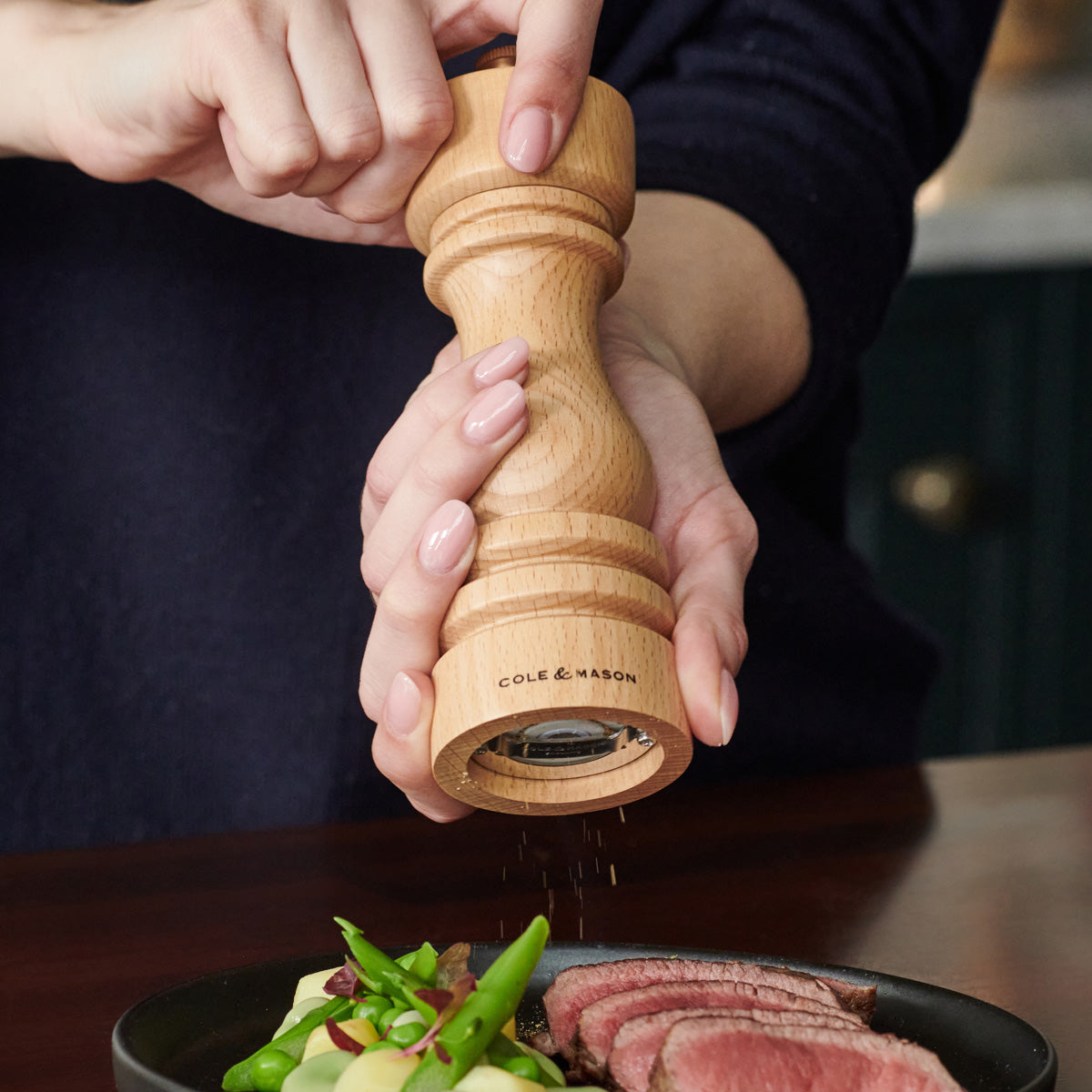 A person uses a wooden Cole & Mason pepper grinder to season a plate of food with sliced meat and vegetables.