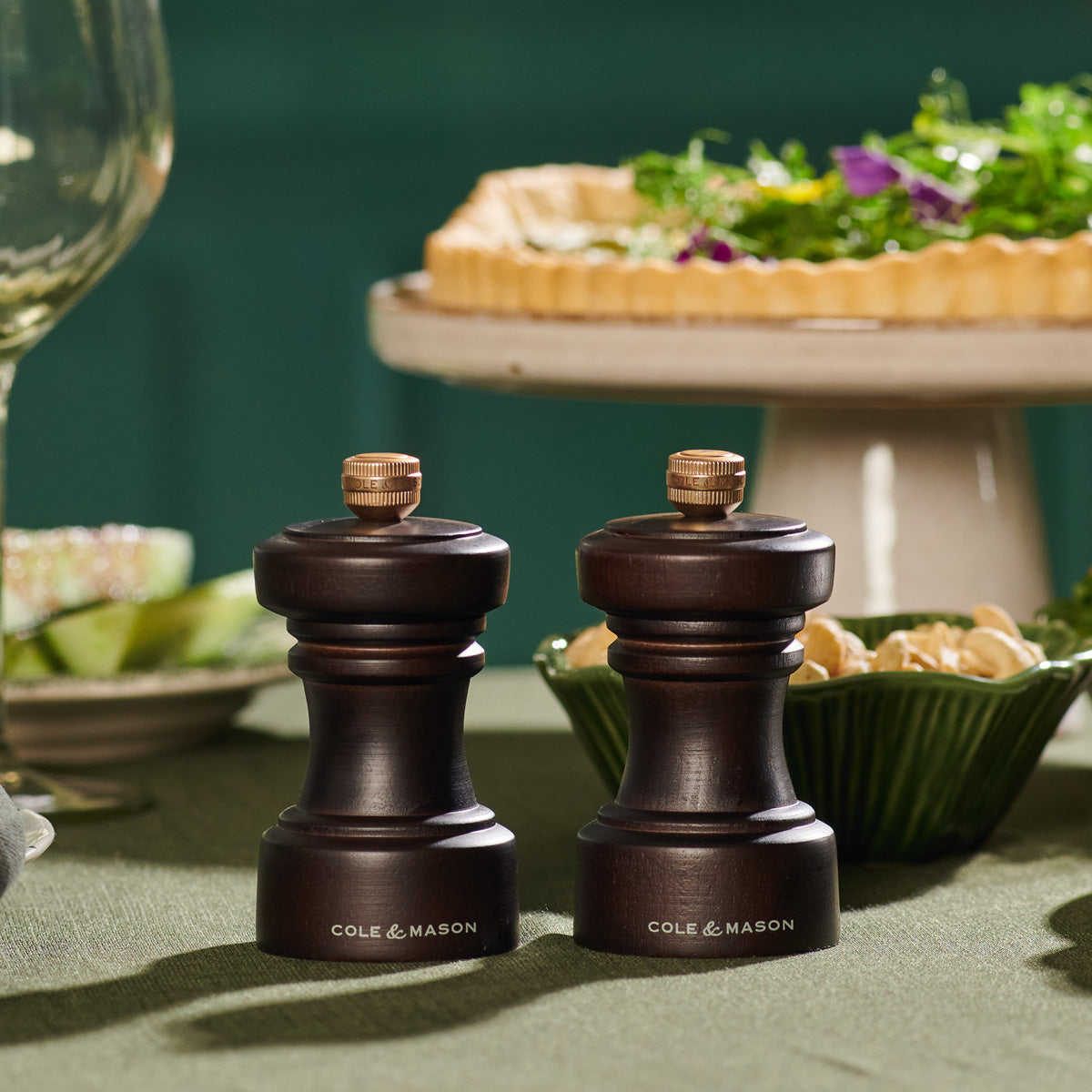 Two dark wooden Cole & Mason salt and pepper grinders sit on a green tablecloth, with a salad, pie, and glassware blurred in the background.