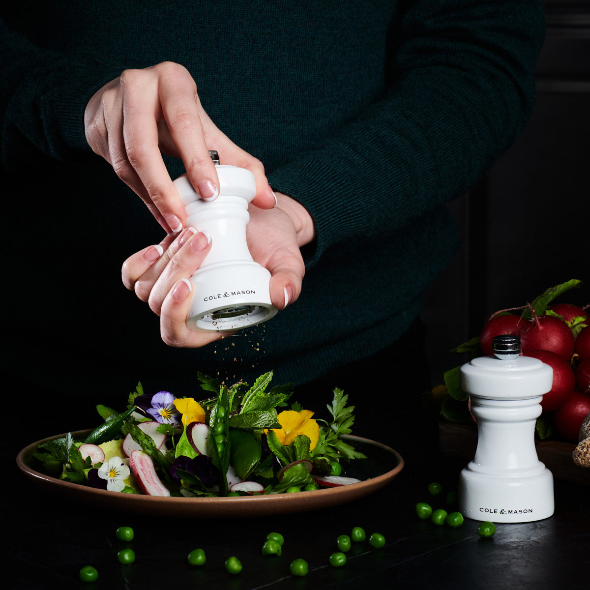 A person grinds pepper from a white pepper mill over a plate of fresh salad with greens and edible flowers; another identical pepper mill sits nearby.