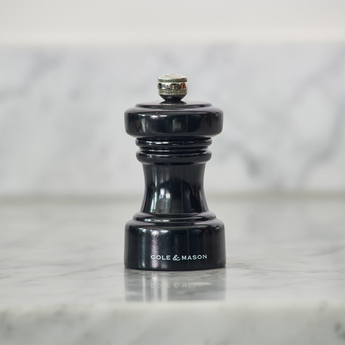 A small black pepper grinder labeled Cole & Mason sits on a light gray marble countertop with a blurred marble backsplash in the background.