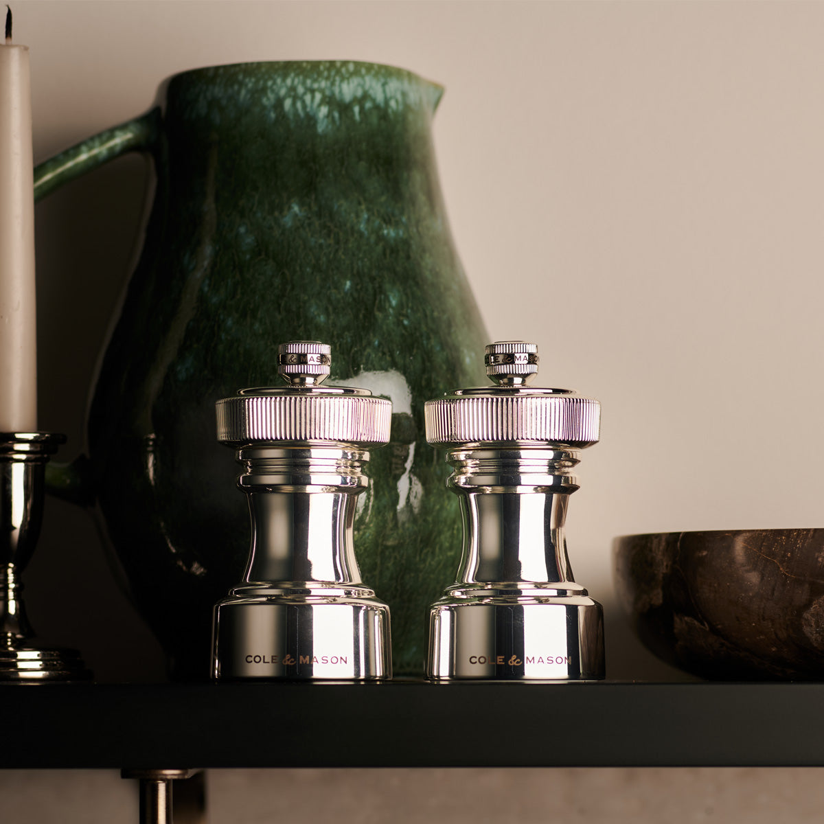 Two metallic salt and pepper grinders with Cole & Mason branding are displayed on a shelf, alongside a green ceramic pitcher, a wooden bowl, and part of a white candle.