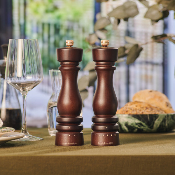 Two dark wooden salt and pepper grinders sit on a green tablecloth beside a wine glass, plates, and a bowl of bread, with blurred greenery and windows in the background.
