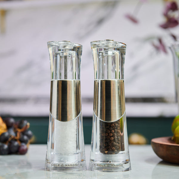 Two transparent grinders, one filled with white salt crystals and the other with black peppercorns, sit side by side on a kitchen countertop. Both have silver accents and are labeled Cole & Mason.