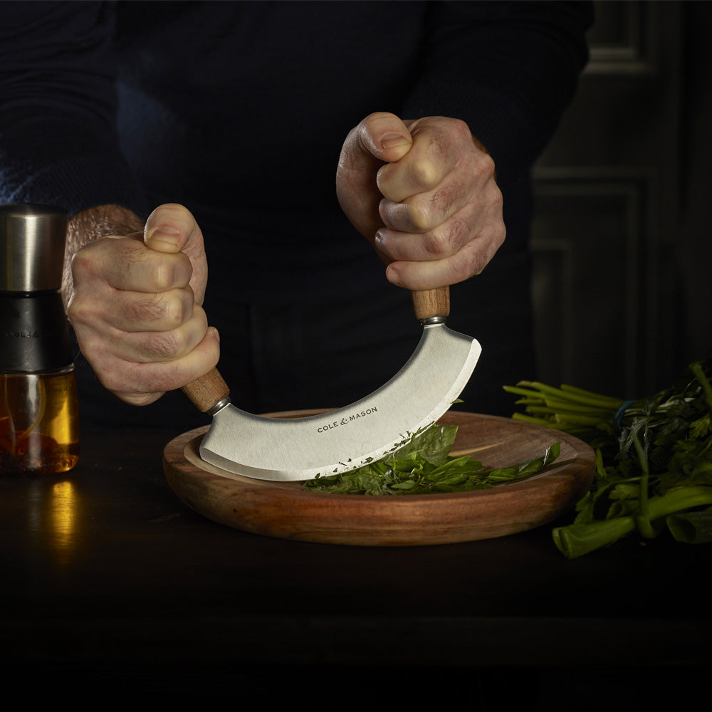 A person uses the Cole & Mason Single Herb Keeper & Chopper Set to cut fresh herbs on a wooden plate, with the herb keeper and other kitchen items visible on the dark surface.