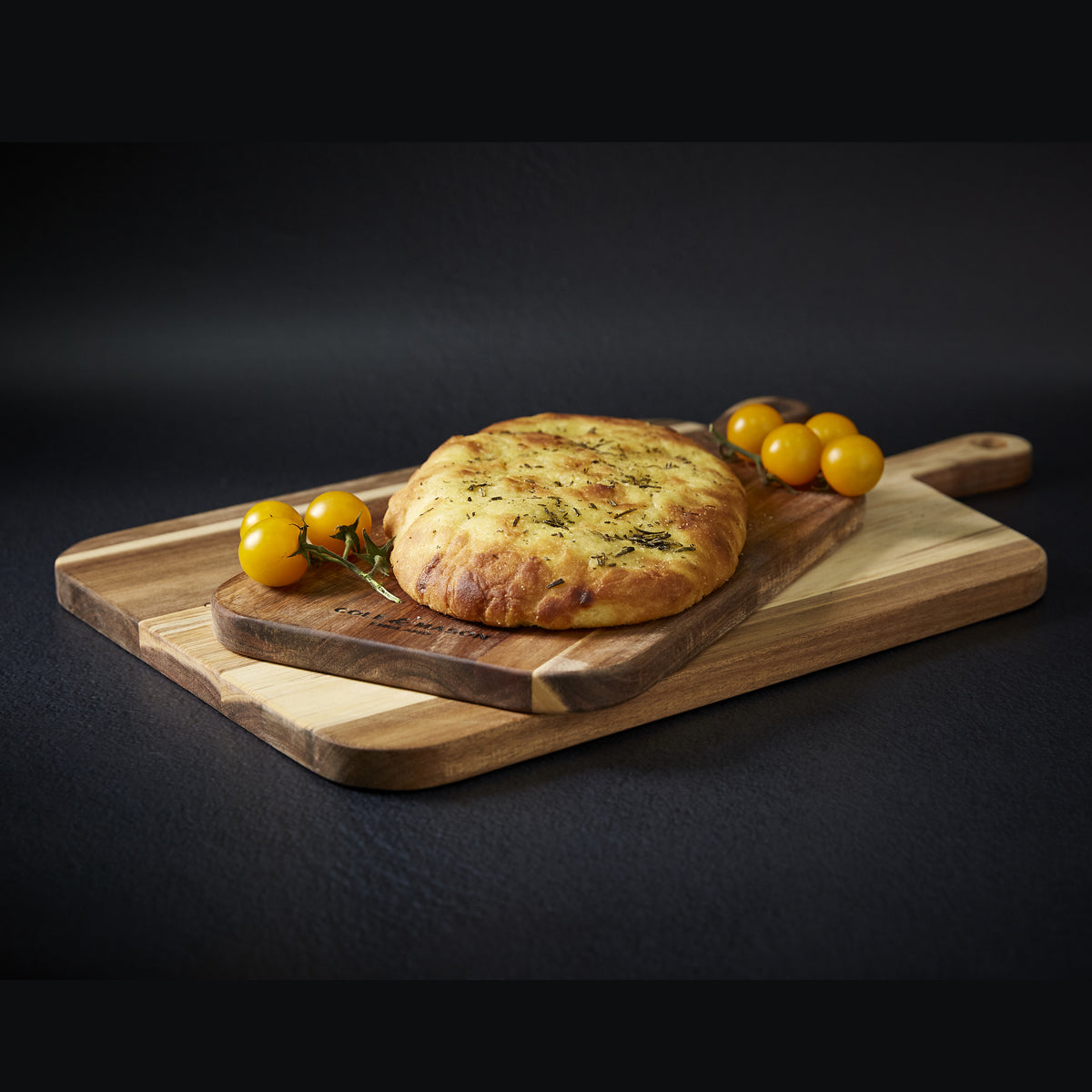 A loaf of focaccia bread sits on a wooden cutting board with a few yellow cherry tomatoes beside it, all placed on a larger cutting board against a dark background.