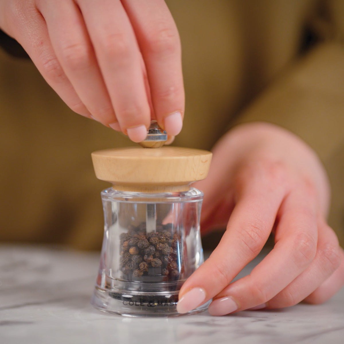 A person’s hands hold and twist a wooden-topped pepper grinder with whole black peppercorns inside, set on a marble countertop.