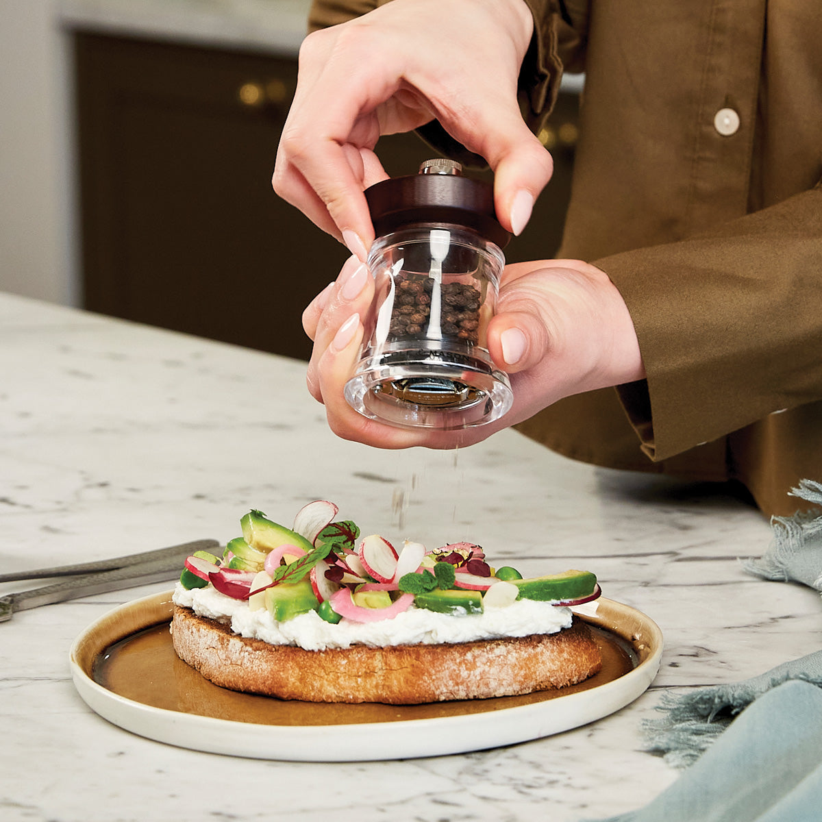 A person sprinkles pepper from a grinder onto a piece of toast topped with ricotta, sliced radishes, and green vegetables on a tan plate. The scene is set on a marble countertop.