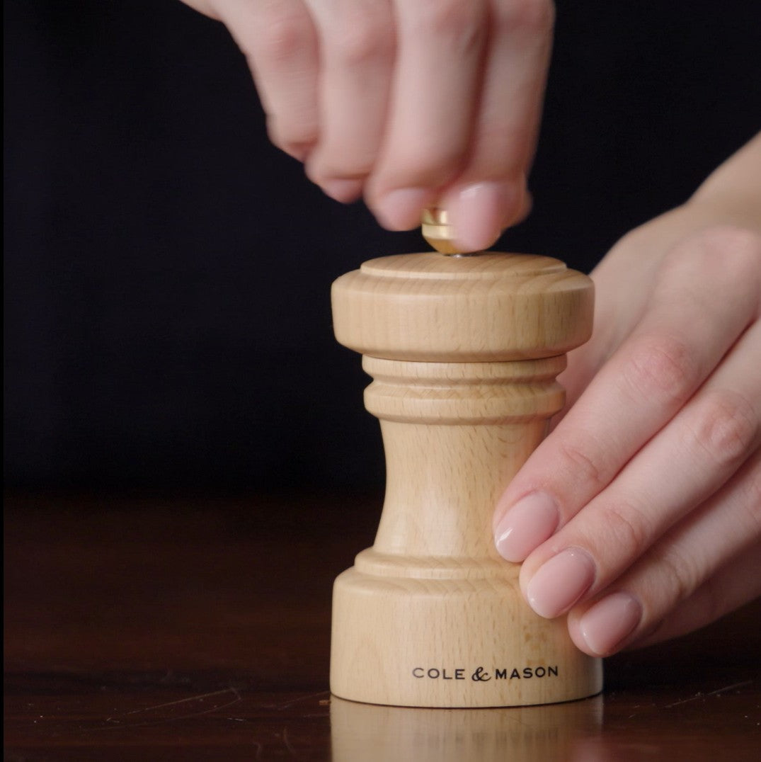 A person’s hands are holding and twisting a wooden Cole & Mason pepper grinder on a dark surface.