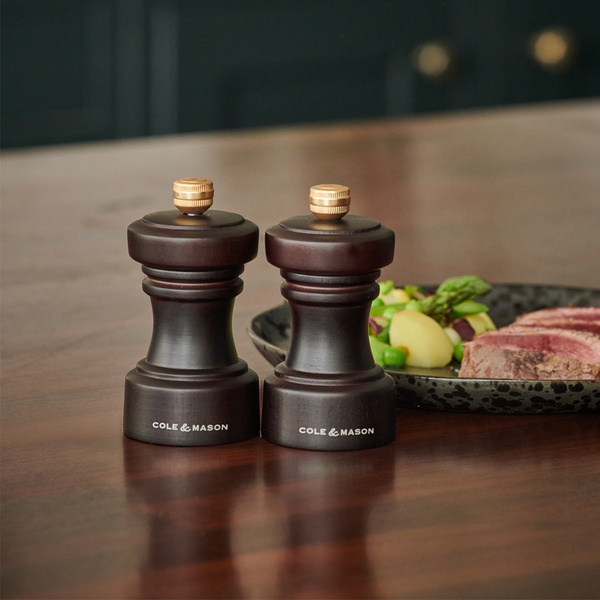 Two dark wooden Cole & Mason salt and pepper grinders are placed on a wooden table next to a plate of sliced steak, potatoes, and vegetables.