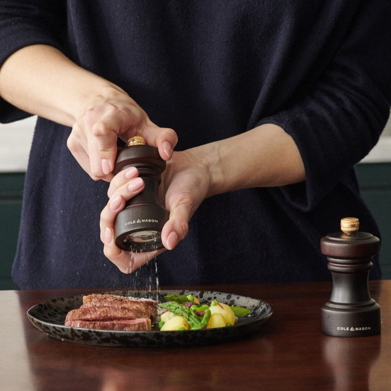 A person grinds pepper over a plate of steak and vegetables using a dark wooden Cole & Mason pepper mill, with a matching salt mill on the table nearby.