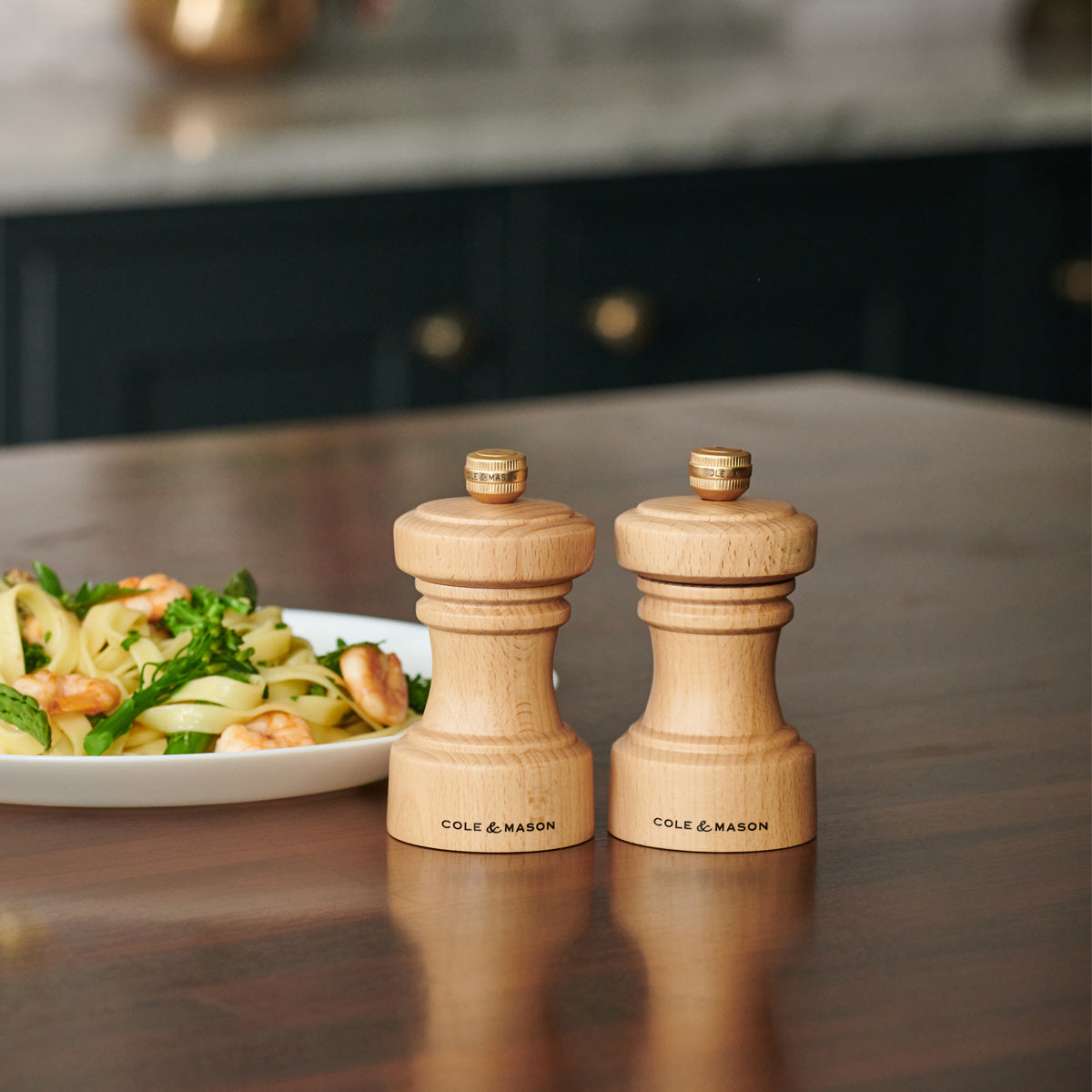 Two wooden Cole & Mason salt and pepper grinders sit on a kitchen counter next to a plate of pasta with greens and nuts. The background includes dark cabinetry and a marble countertop.