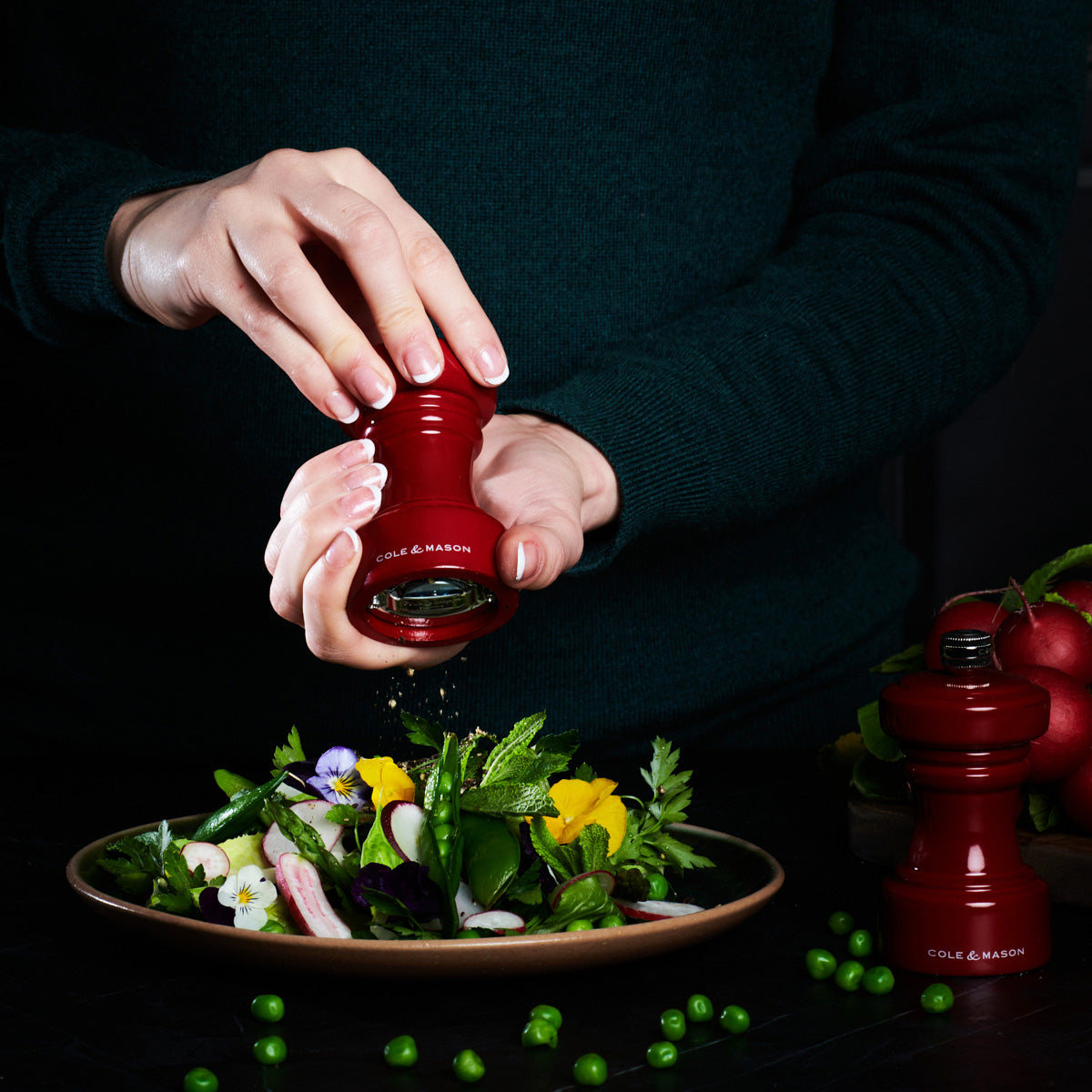 A person in a dark green sweater grinds pepper from a red pepper grinder over a fresh salad on a brown plate, with another red grinder and green peas nearby on a dark surface.
