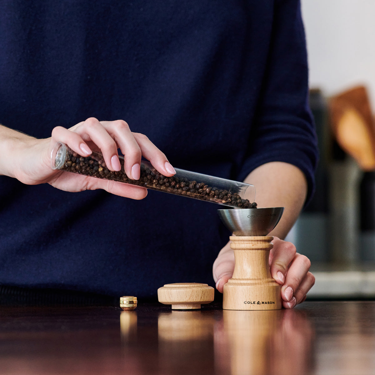 A person pours whole black peppercorns from a glass tube into a wooden pepper grinder with a metal funnel, on a dark countertop.