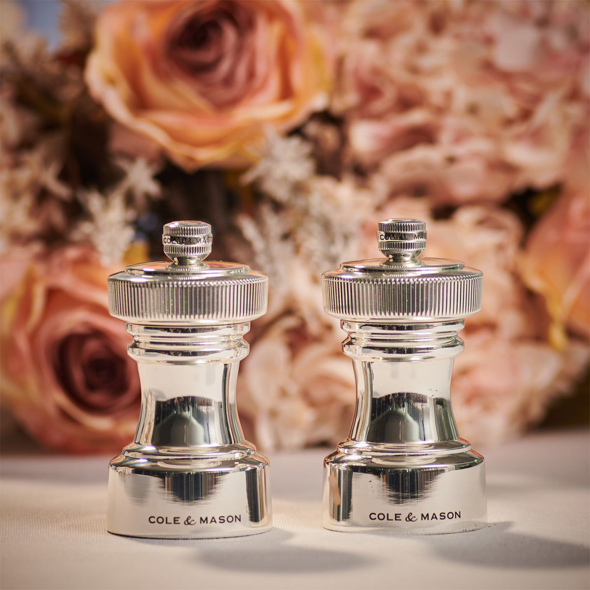 Two silver salt and pepper grinders labeled COLE & MASON stand side by side in front of a blurred background of pink and peach roses.