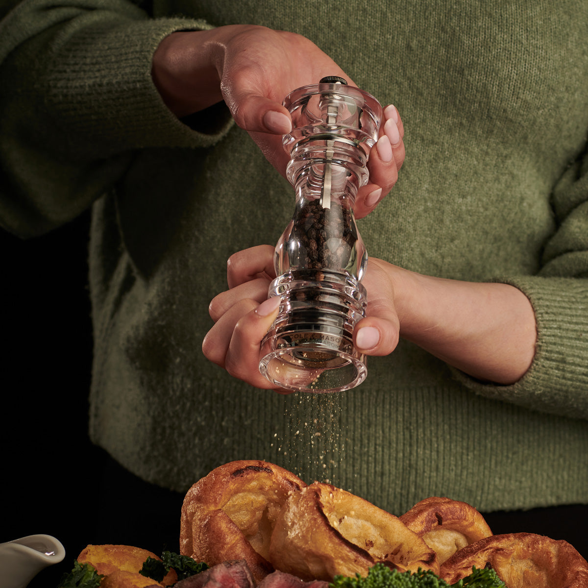 A person wearing a green sweater uses a clear pepper grinder to season food on a plate below, which includes Yorkshire puddings and broccoli.