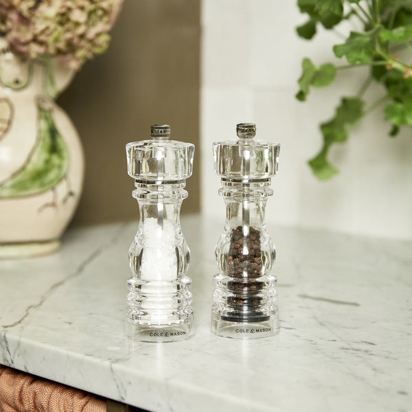 A pair of clear Cole & Mason salt and pepper grinders sit side by side on a marble kitchen countertop, with greenery and a decorative ceramic vessel in the background.