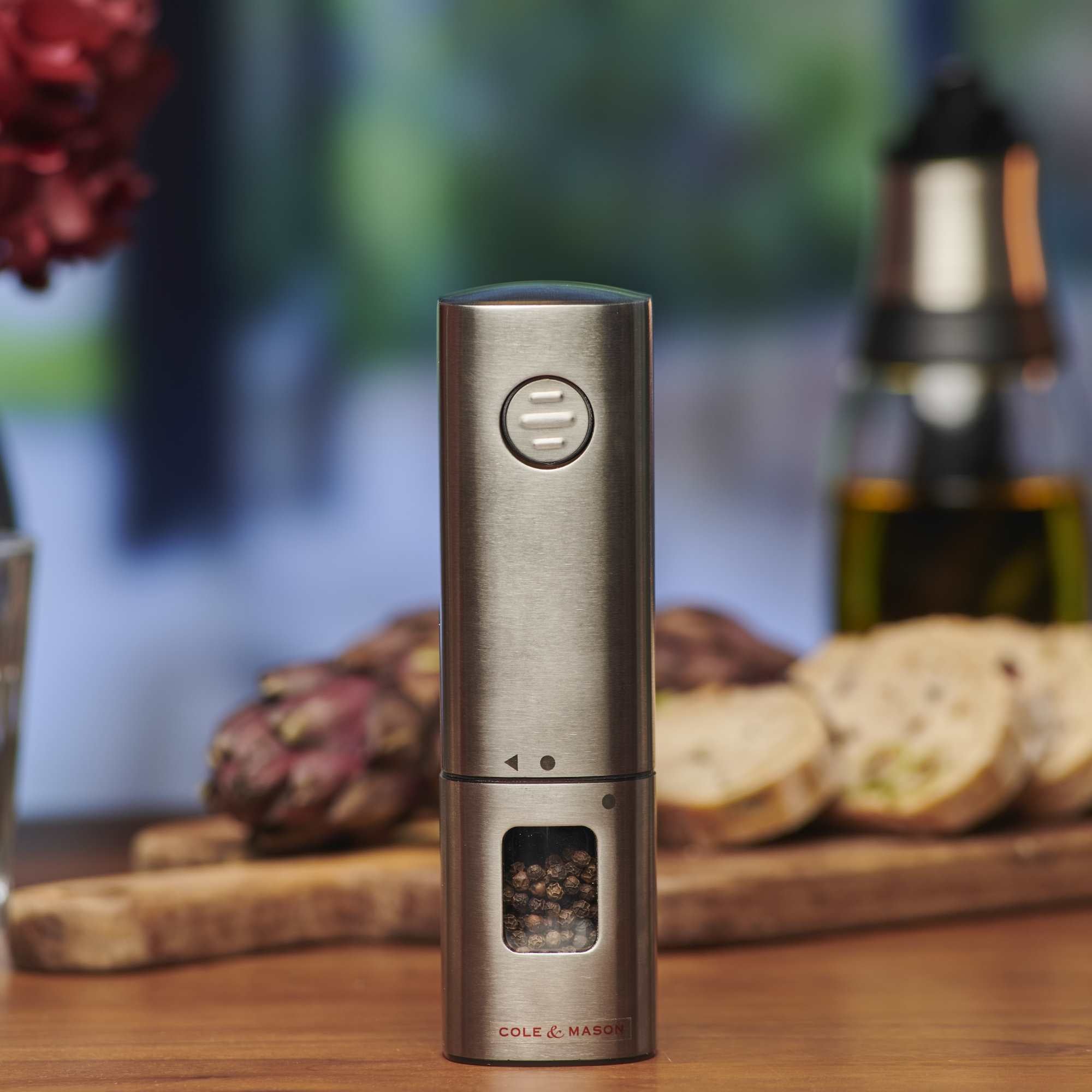 A stainless steel electric pepper grinder with visible peppercorns, placed on a wooden table. Sliced bread, artichokes, and a bottle of oil are blurred in the background.