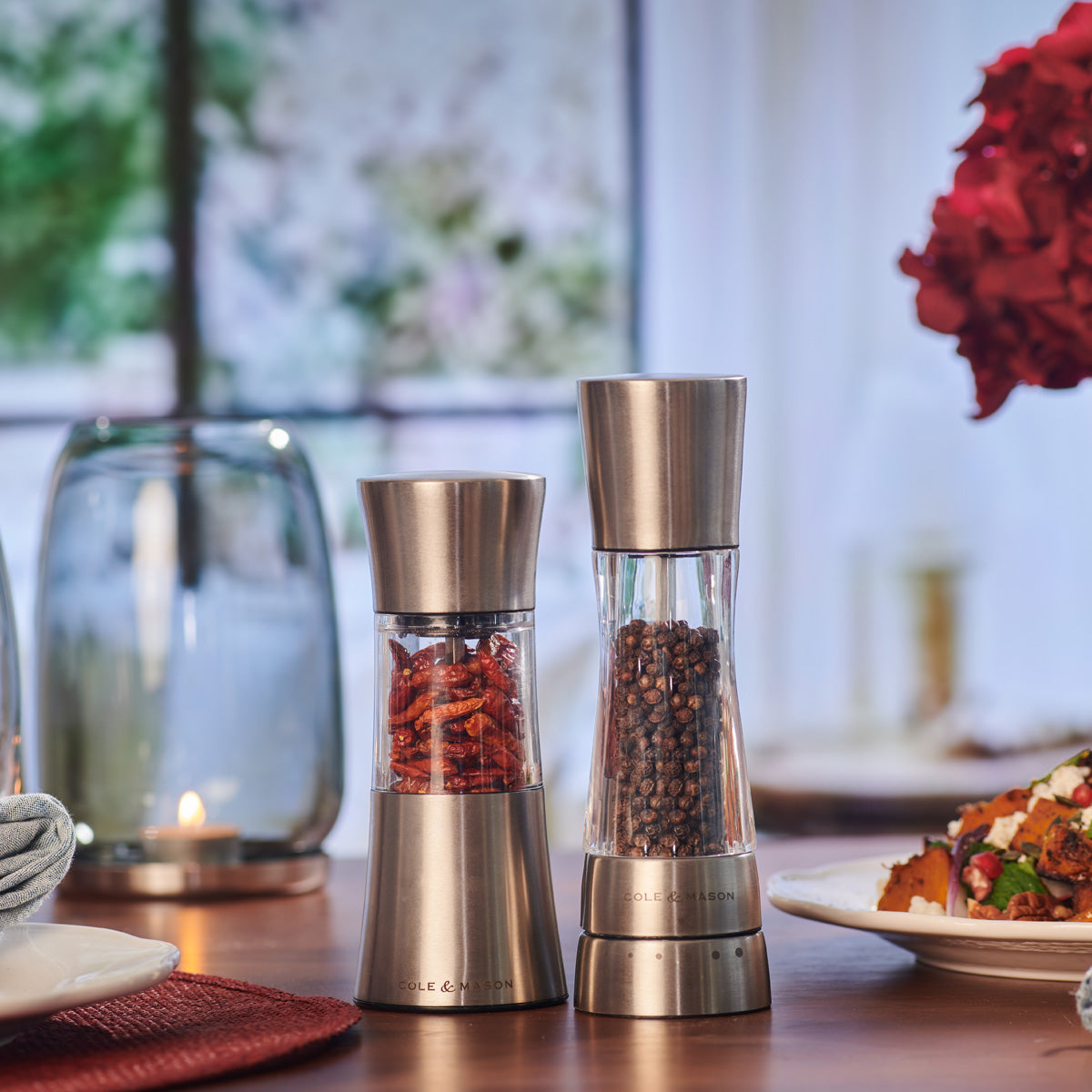 Two stainless steel and glass grinders filled with dried chili flakes and black peppercorns sit on a table set for a meal, with dishes and a candle in the background.