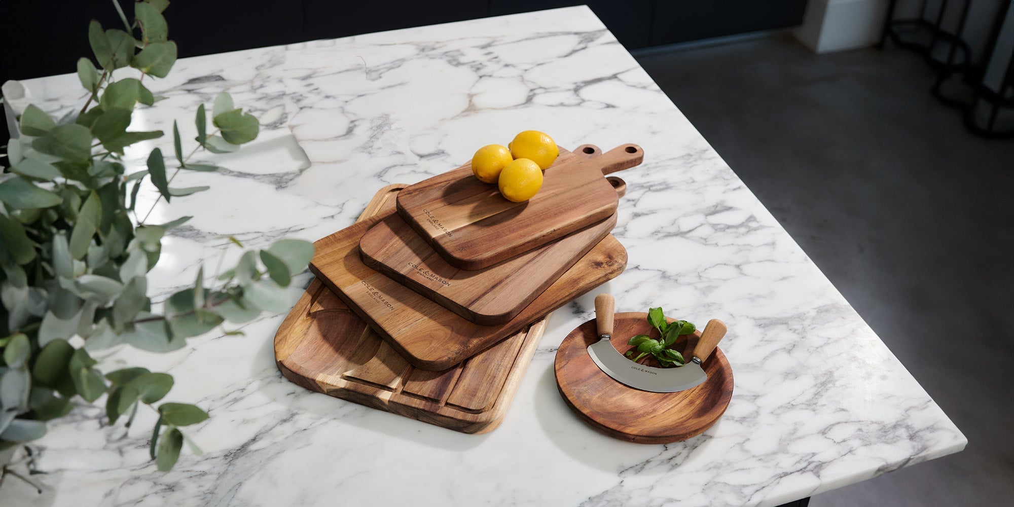 A stack of wooden cutting boards and three yellow lemons on a marble countertop, with a mezzaluna knife on a small wooden plate and green leaves in the foreground.