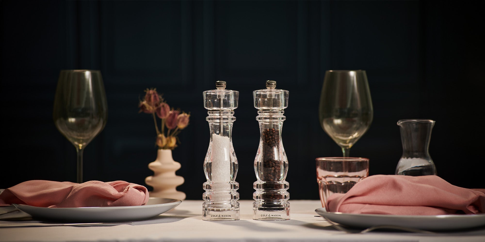 A neatly set dining table with two plates, pink napkins, empty wine glasses, a vase with dried flowers, a glass pitcher, and clear salt and pepper mills at the center. The background is dark and elegant.