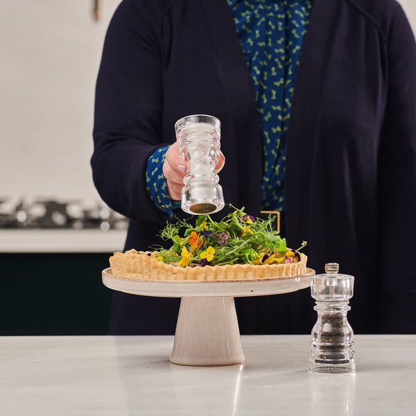 A person sprinkles pepper from the Cole & Mason London Acrylic Salt Shaker & Pepper Mill Set onto a tart with greens and edible flowers on a pedestal stand, while the modern set completes the stylish tableware display.
