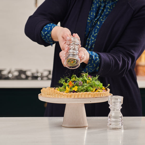 A person grinds fresh pepper from the Cole & Mason London Acrylic Salt Shaker & Pepper Mill Set onto a vegetable tart with greens and edible flowers, displayed on a cake stand among stylish tableware on the kitchen counter.