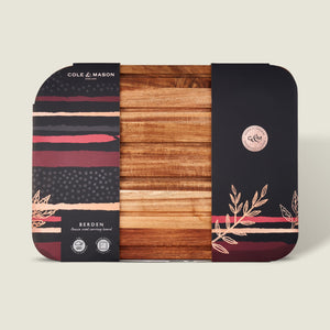 The rectangular Berden Acacia Carving Board by Cole & Mason comes partially wrapped in stylish packaging with black, red, and beige foliage patterns—an elegant addition to your kitchen.