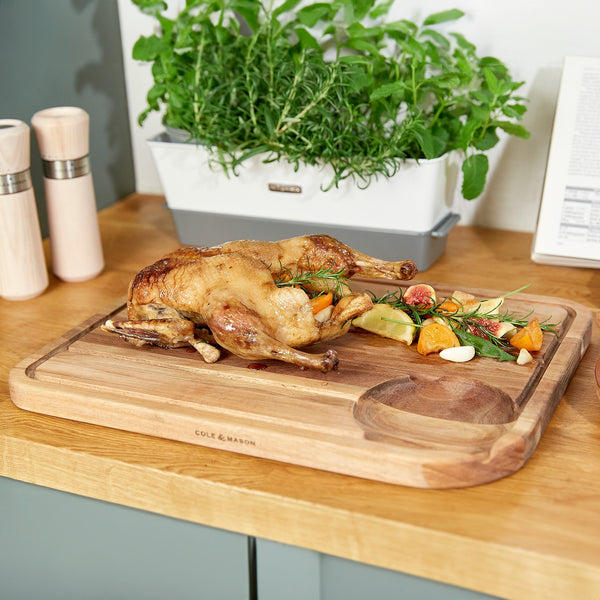 A whole roasted duck rests on the Cole & Mason Berden Acacia Carving Board with juice grooves, surrounded by sliced vegetables and figs, while fresh herb plants in the background add to the kitchen’s appeal.