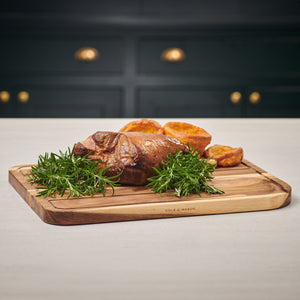 A crispy roast rests on the Cole & Mason Berden Acacia Carving Board, garnished with rosemary and Yorkshire puddings. Juice grooves catch drippings, complementing a kitchen with dark drawers and brass handles.
