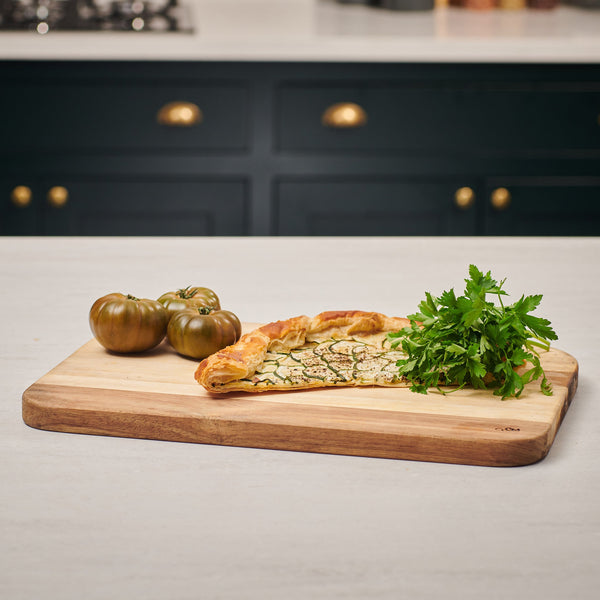 The Cole & Mason Berden Acacia Carving Board on the kitchen counter holds a slice of vegetable galette, parsley, and heirloom tomatoes. Juice grooves offer function while dark cabinets and a stove add style to the background.