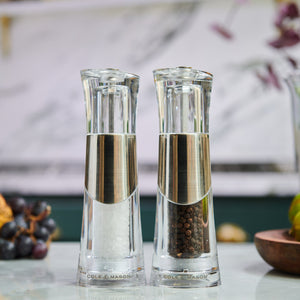 The Cole & Mason Bobbi Salt & Pepper Mill Set features two clear grinders with silver accents and a durable mechanism, displayed side by side—one filled with coarse salt, the other with black peppercorns against a softly blurred background.