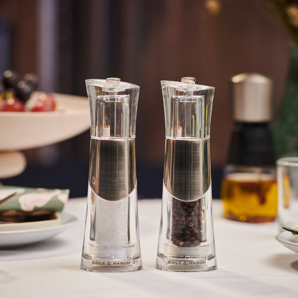 The Cole & Mason Bobbi Salt & Pepper Mill Set, with a clear modern design and silver accents, sits side by side on the table, showcasing a durable grinding mechanism among dishes in a softly lit dining space.