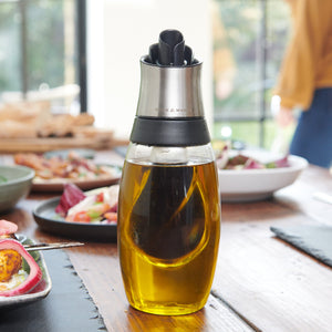 The Cole & Mason Bristol Duo Oil & Vinegar Pourer, featuring a dual-chamber design for mess-free dispensing, sits on a wooden dining table with plates and salad, while a blurred person stands in the background.