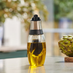 The Cole & Mason Bristol Duo Oil & Vinegar Pourer, made of glass and metal with a pour spout for mess-free dispensing, sits on a kitchen counter. A blurred artichoke and greenery can be seen in the background.