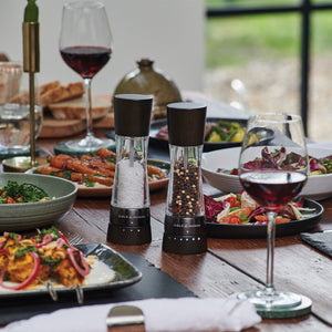 A wooden table set for a meal with plates of colorful food, two glasses of red wine, and salt and pepper grinders at the center. Bread and candles are also visible in the background.