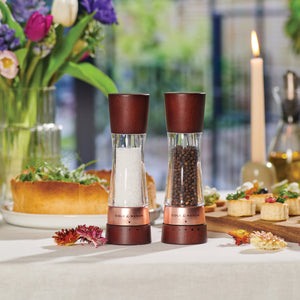 A set table with a salt and pepper grinder set in the foreground, surrounded by flowers, a lit candle, bread, and assorted appetizers, creating an elegant dining atmosphere.