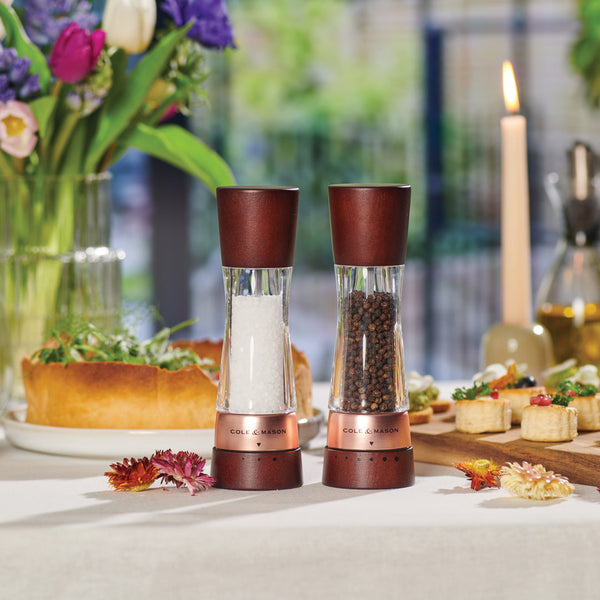 A set table with a salt and pepper grinder set in the foreground, surrounded by flowers, a lit candle, bread, and assorted appetizers, creating an elegant dining atmosphere.