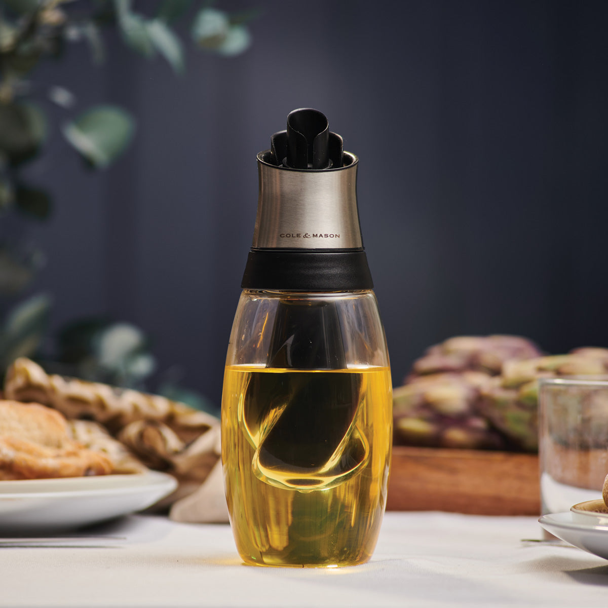 Close-up of a glass oil dispenser with a stainless steel top, filled with yellow oil, placed on a table set with plates, food, and blurred greenery in the background.