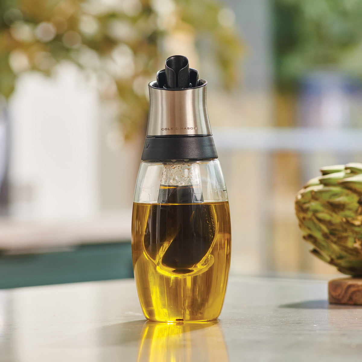 A glass bottle filled with golden yellow oil, topped with a stainless steel and black pour spout, sits on a countertop with blurred greenery and part of an artichoke in the background.