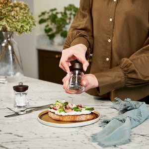 A person in a brown shirt uses a pepper grinder to season an open-faced sandwich with vegetables on a plate, on a marble kitchen counter with a napkin, fork, and knife nearby.